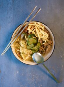 Bowl of Kuksi cold noodle soup with colorful toppings like stir-fried beef, cabbage salad, cucumber, daikon, egg noodles, and red pepper oil.