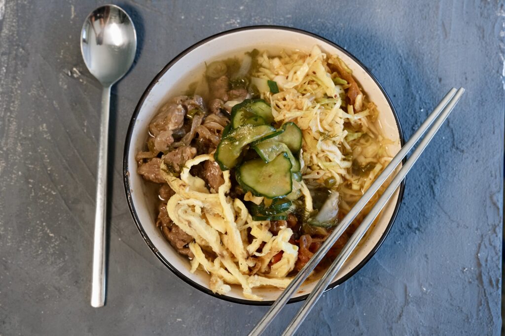 Bowl of Kuksi cold noodle soup with colorful toppings like stir-fried beef, cabbage salad, cucumber, daikon, egg noodles, and red pepper oil.
