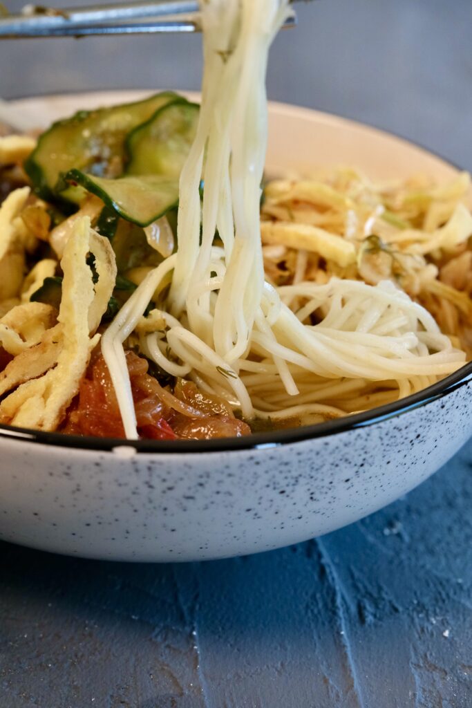 Bowl of Kuksi cold noodle soup with colorful toppings like stir-fried beef, cabbage salad, cucumber, daikon, egg noodles, and red pepper oil.