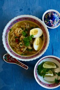 A bowl of soto ayam – Indonesian chicken soup with shredded chicken, rice noodles, boiled egg, fresh herbs, and a golden turmeric broth.