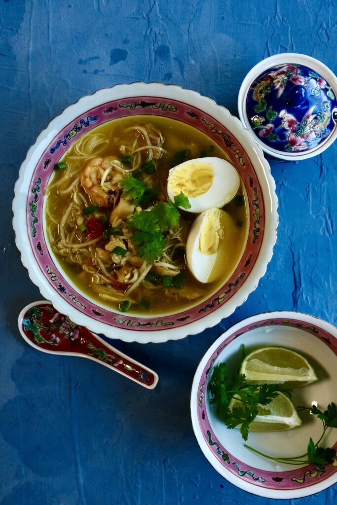 A bowl of soto ayam – Indonesian chicken soup with shredded chicken, rice noodles, boiled egg, fresh herbs, and a golden turmeric broth.