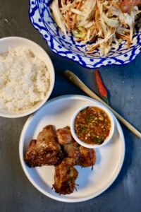Juicy Thai-style Gai Yang chicken, grilled to golden perfection, served with fresh herbs and dipping sauce on a rustic plate.
